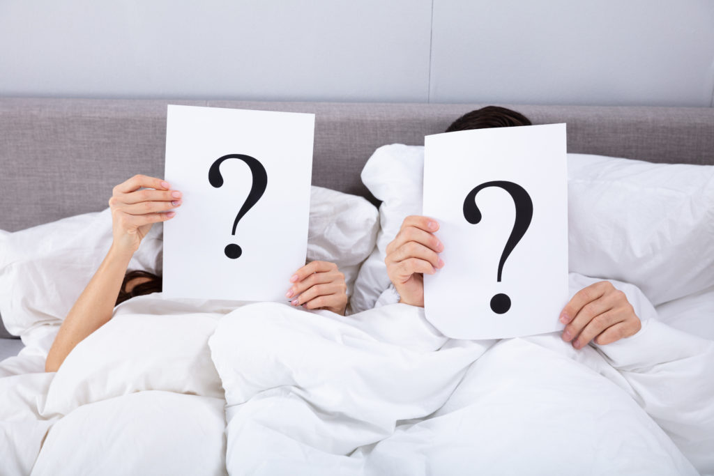 Couple Lying On Bed Holding Question Mark Sign In Front Of Their Face